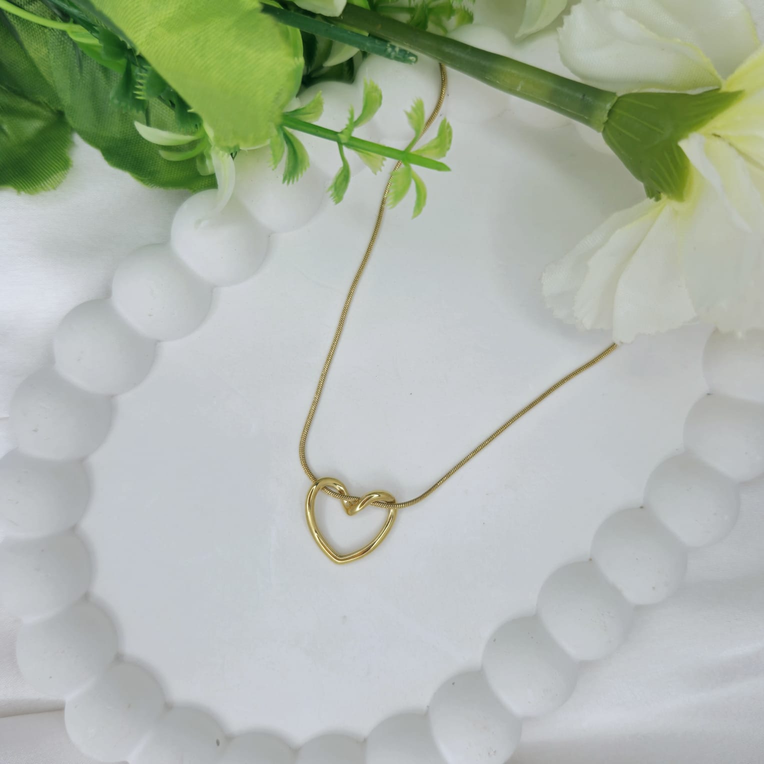 Gold heart-shaped necklace on a white textured surface with green leaves in the background