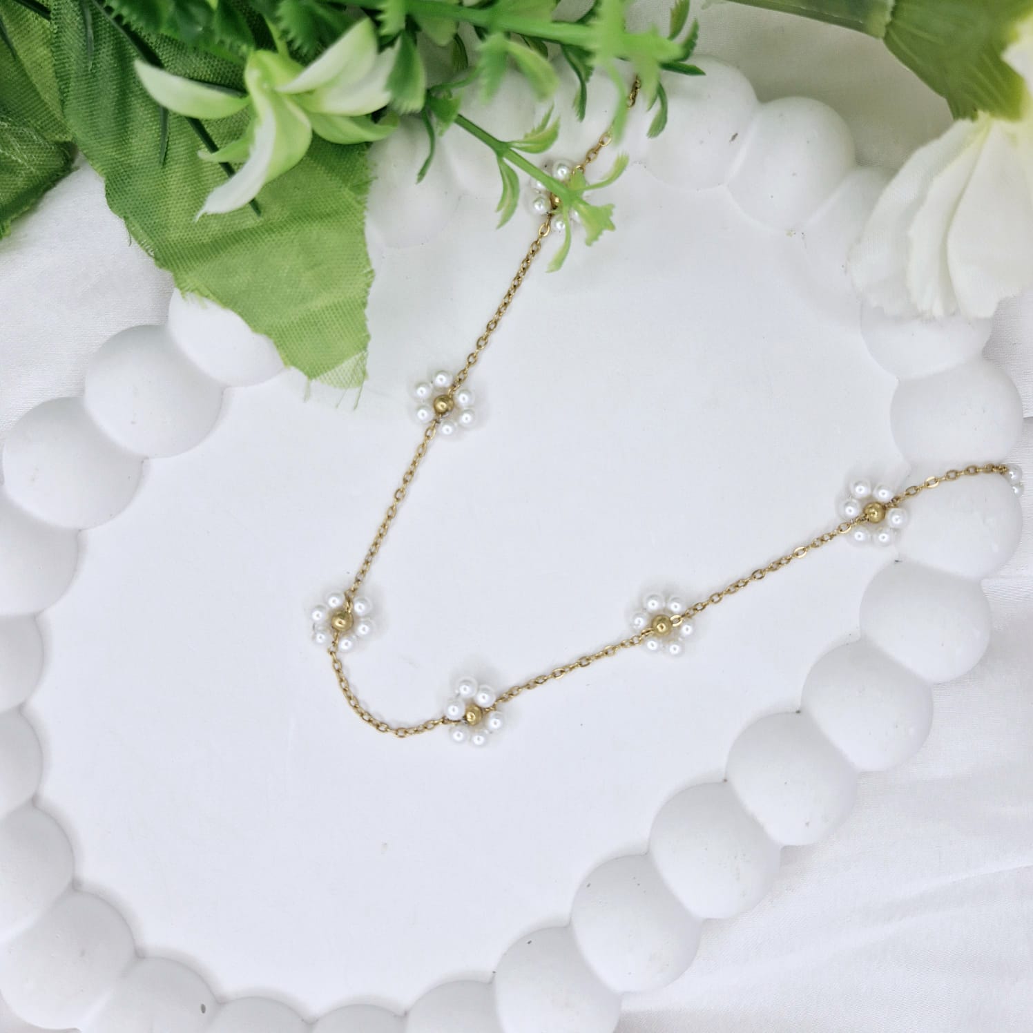 White beaded necklace with gold chain on a white surface with green leaves in the background