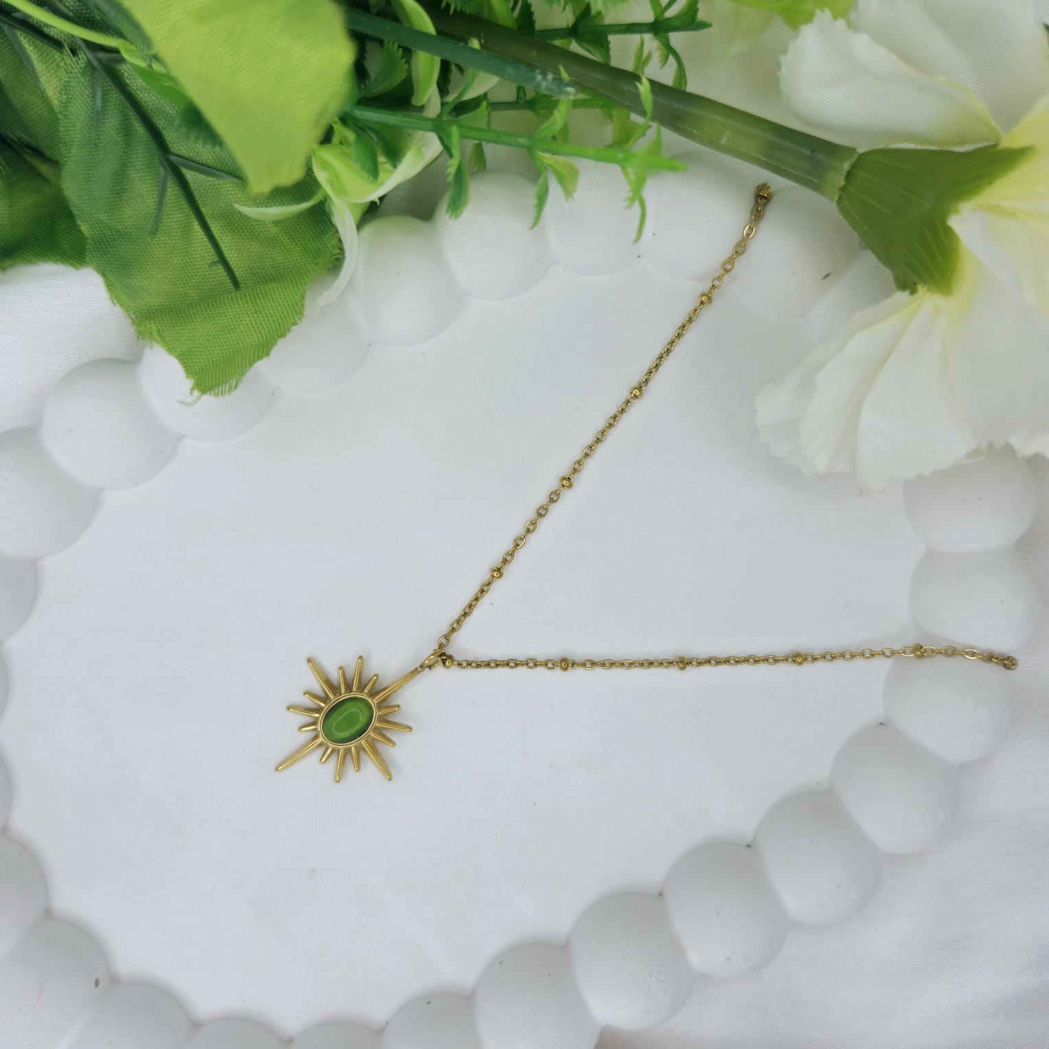 Gold necklace with a green pendant on a white surface with green leaves and flowers.