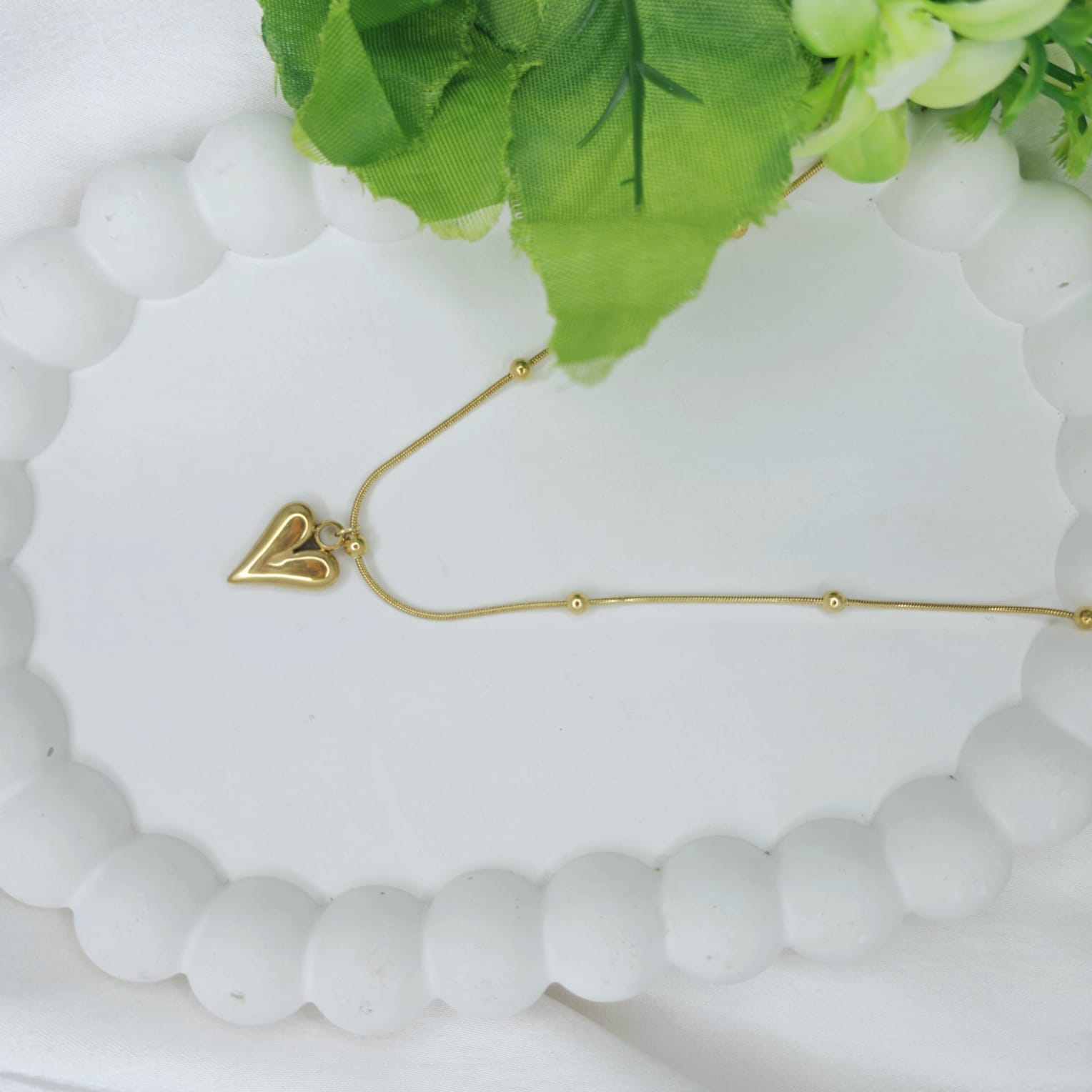 Gold necklace with a heart pendant on a white stone surface with green leaves.