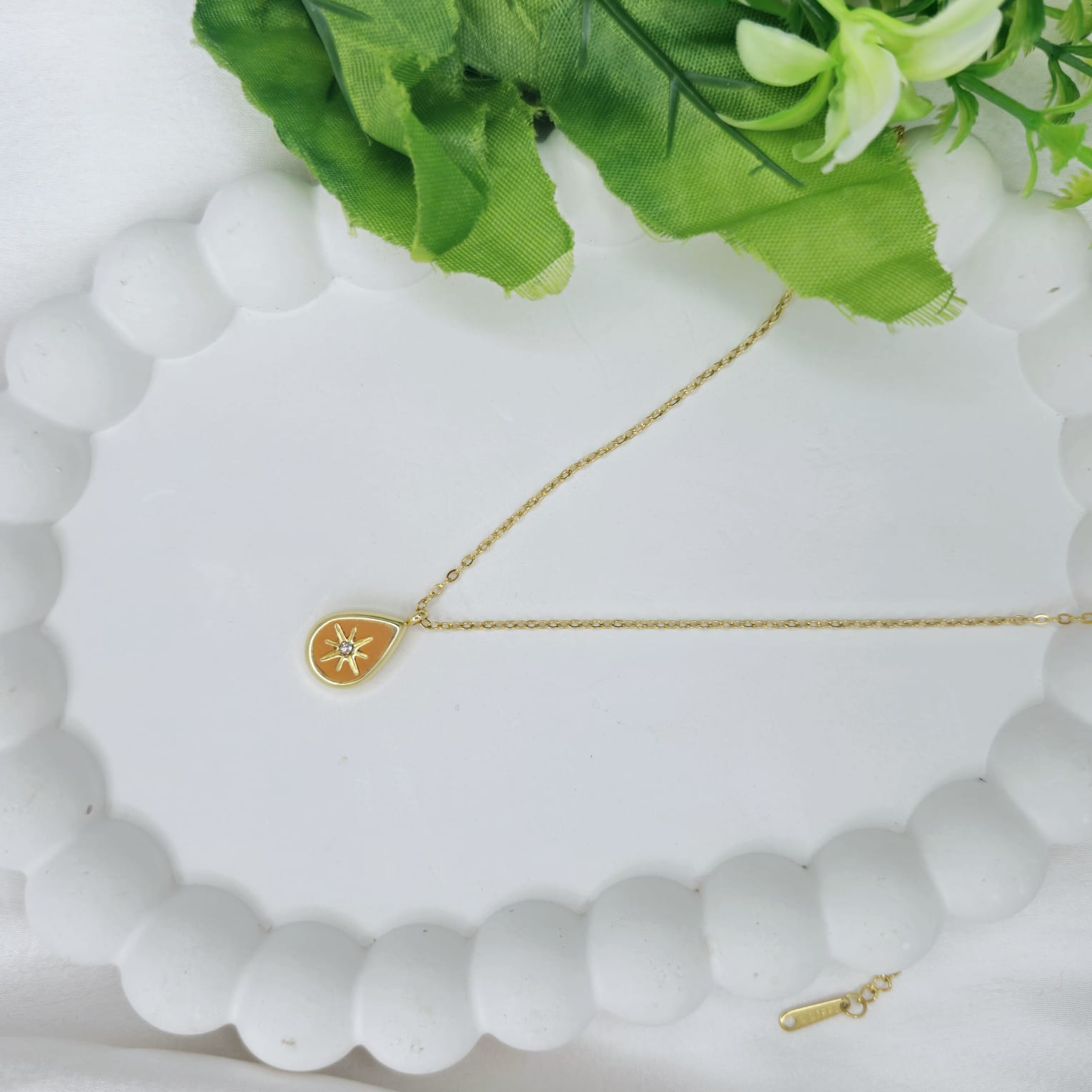 Gold necklace with a heart-shaped pendant on a white surface with green leaves in the background