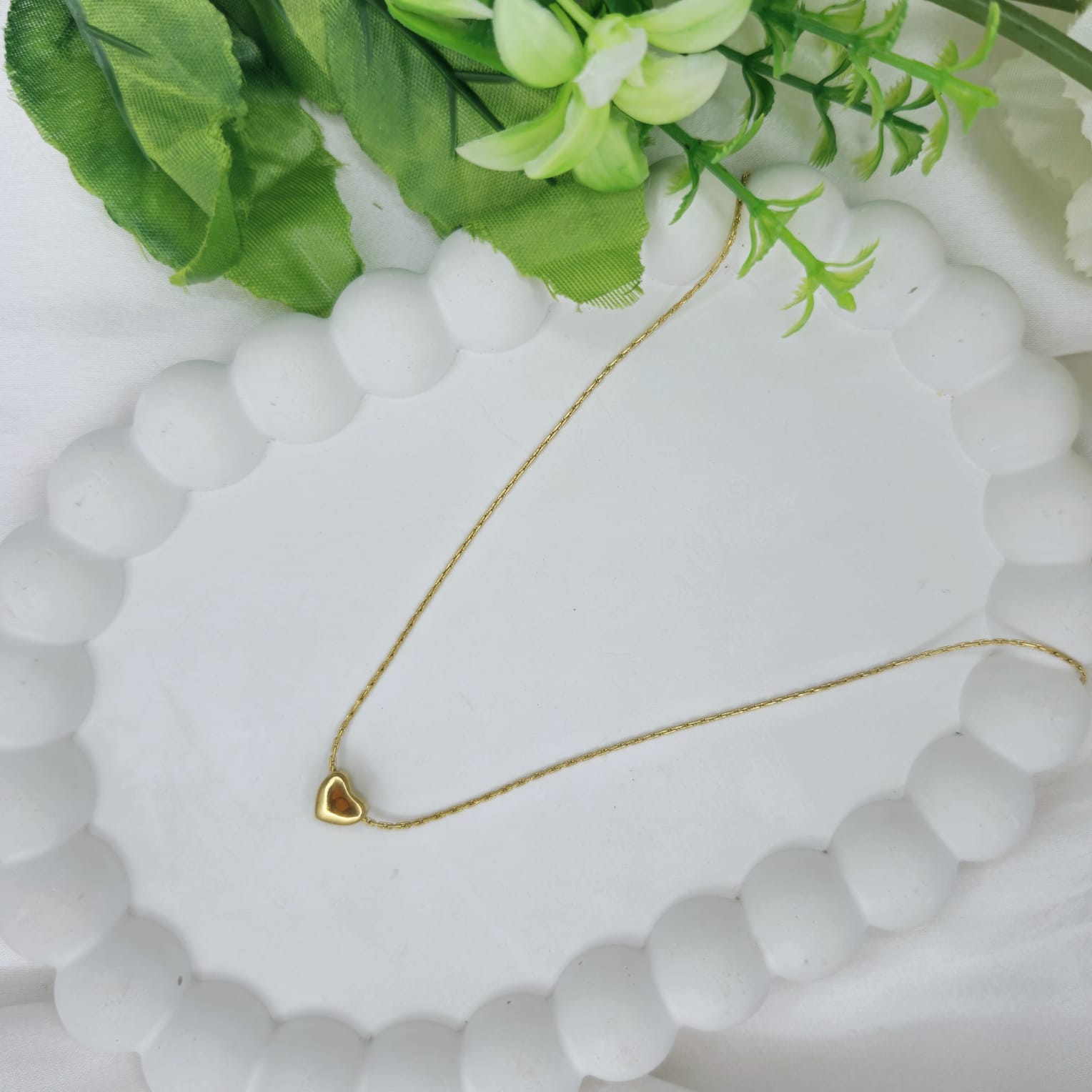 Gold necklace with a heart pendant on a white stone surface with green leaves in the background